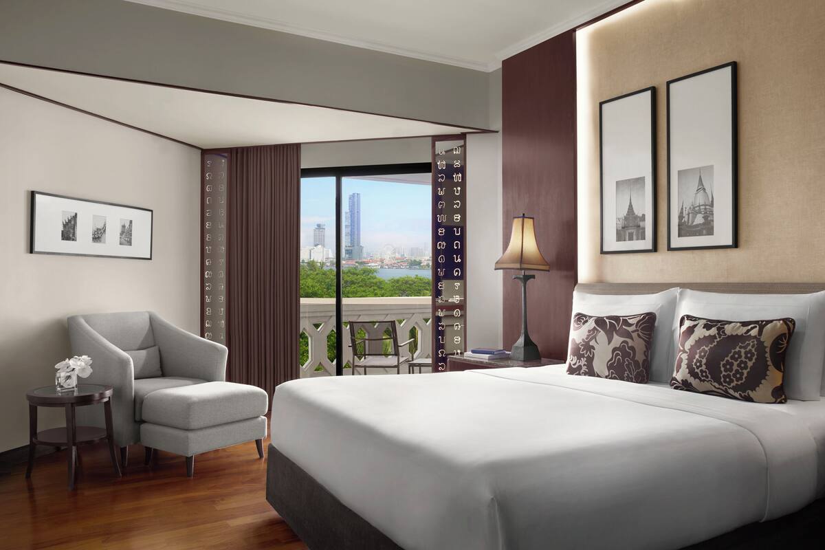 A modern hotel room with a large bed, two bedside lamps, a chair, a small table with a vase, and framed pictures on the wall.