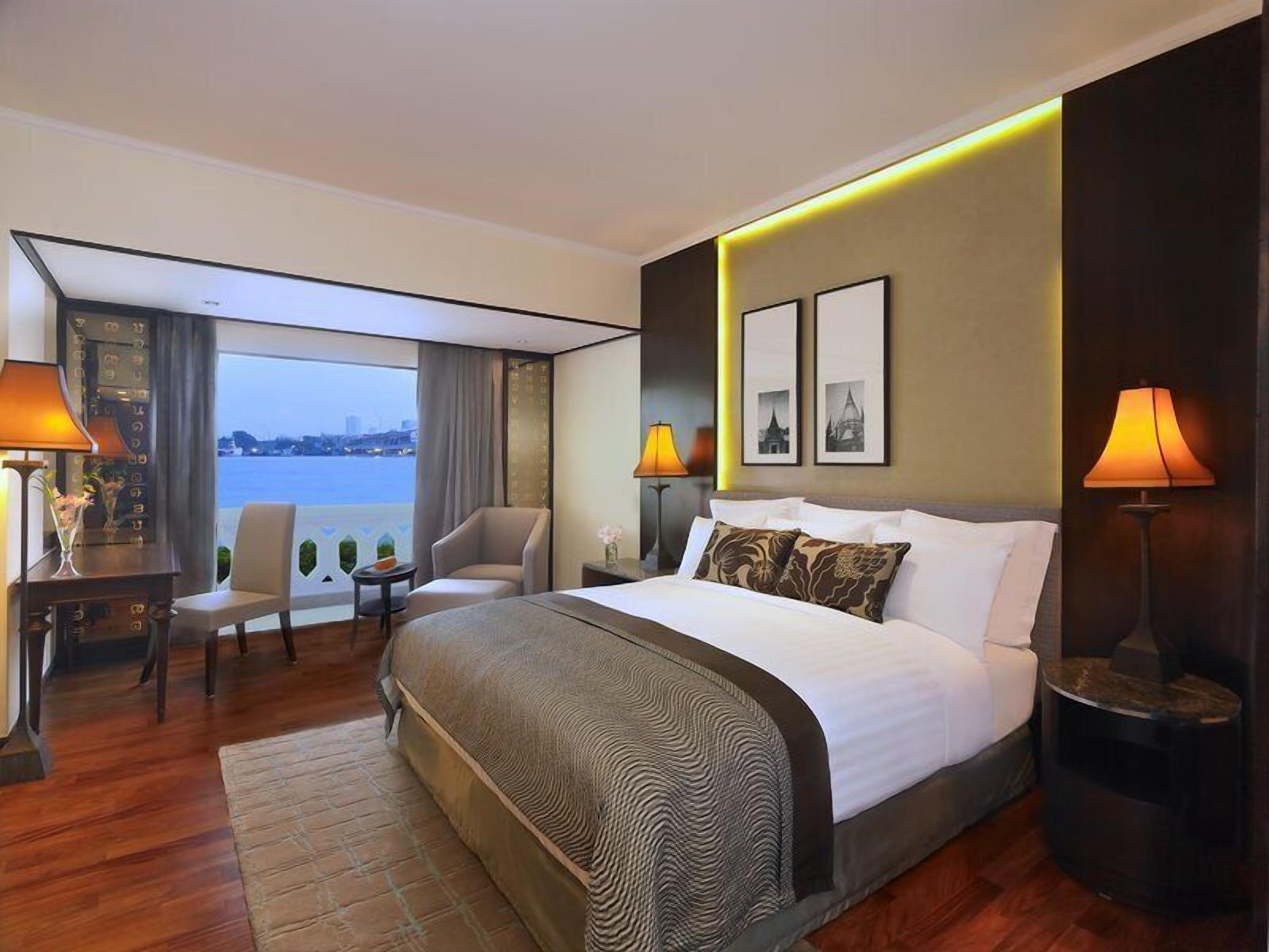 A hotel room with a large bed, two bedside tables, a desk, a chair, and a view of the city.