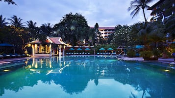 Una gran piscina cristalina rodeada de palmeras y sillas de descanso, con un edificio al fondo.