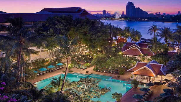 Un resort con una piscina grande rodeada de palmeras y sillas de descanso, con el horizonte de una ciudad al atardecer.