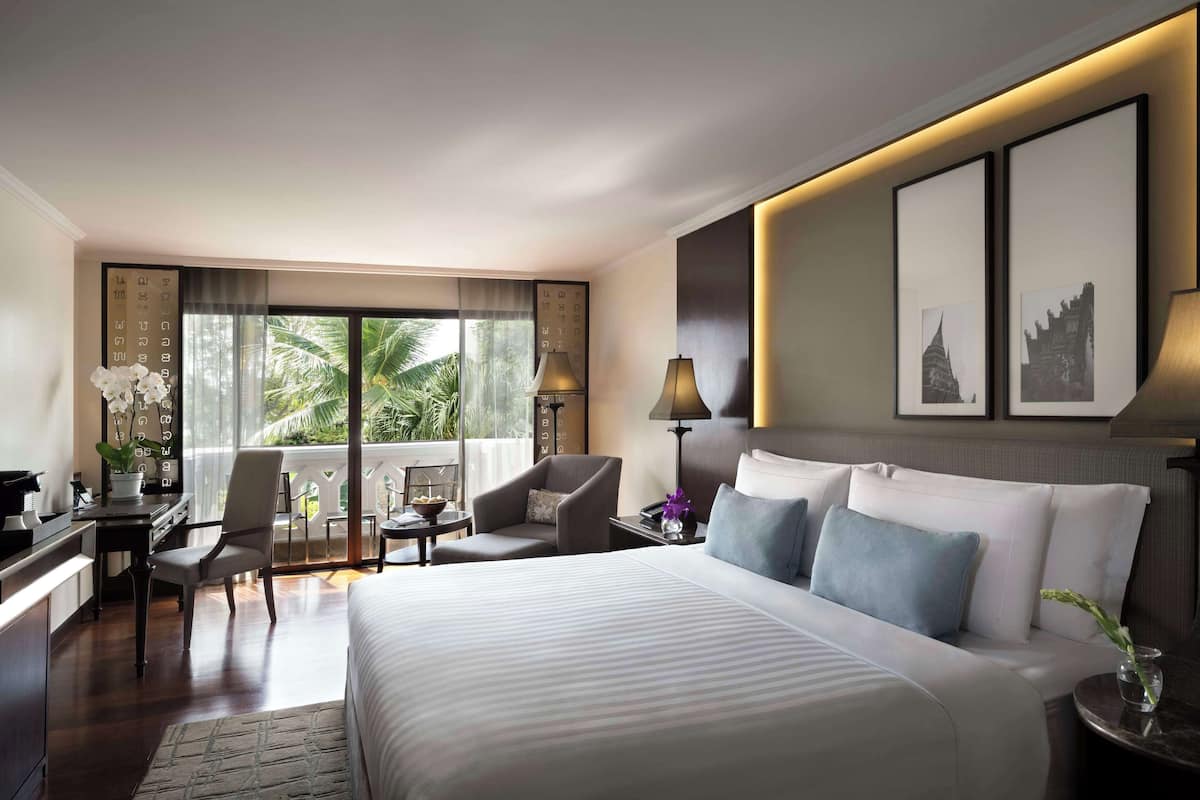 A hotel room with a large bed, a desk, a chair, a small table, and a balcony with a view of palm trees.