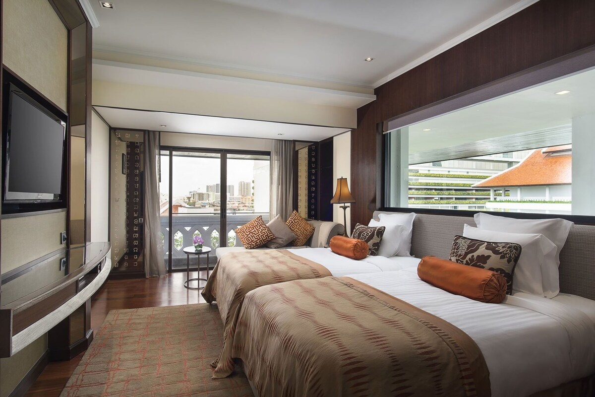 A hotel room with two beds, a TV, a balcony with a city view, and a small table with a lamp.