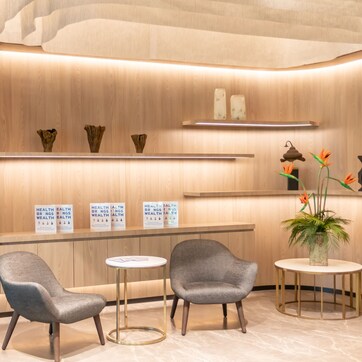 A modern reception area with wooden paneling, two grey upholstered chairs, a small round table, and a potted plant with orange flowers.