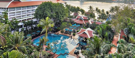 Un hotel con una piscina grande rodeada de palmeras y sombrillas, una noria al fondo y un río.