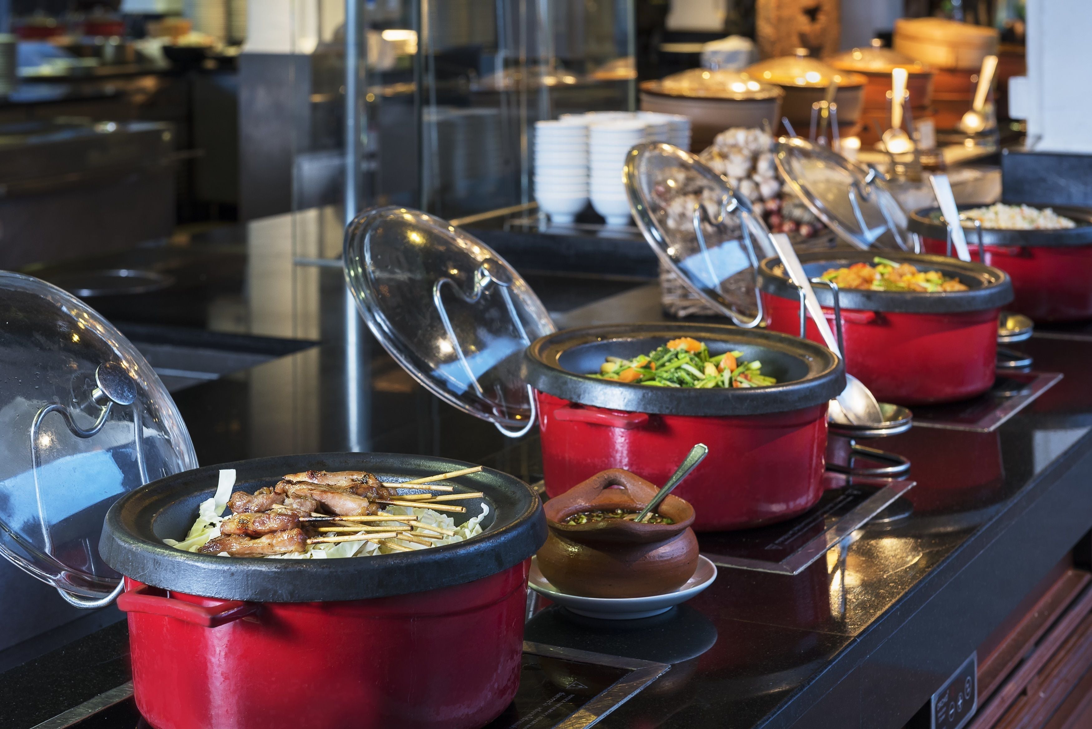 Un bufé con varios platos en ollas rojas, incluyendo una olla con carne ensartada y otra con salsa marrón.
