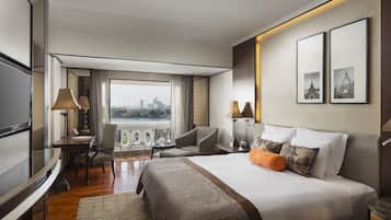 Habitación de hotel con una cama grande, un escritorio, una silla y un televisor. Se ve una vista panorámica de la ciudad a través de la ventana.