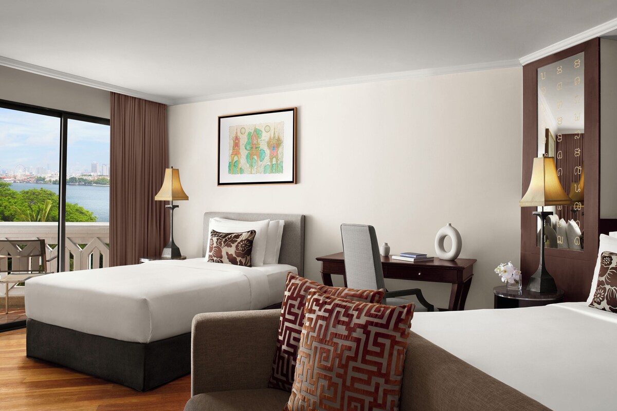 A hotel room with two beds, a desk, a chair, a lamp, and a large window with a city view.