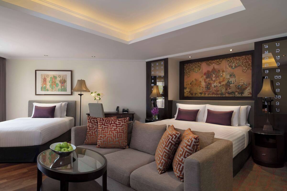 A hotel room with two single beds, a sofa, a glass coffee table, and a large decorative painting on the wall.