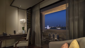 Habitación de hotel moderna con escritorio, sillas, sofá y una amplia ventana con vistas a un paisaje urbano nocturno.