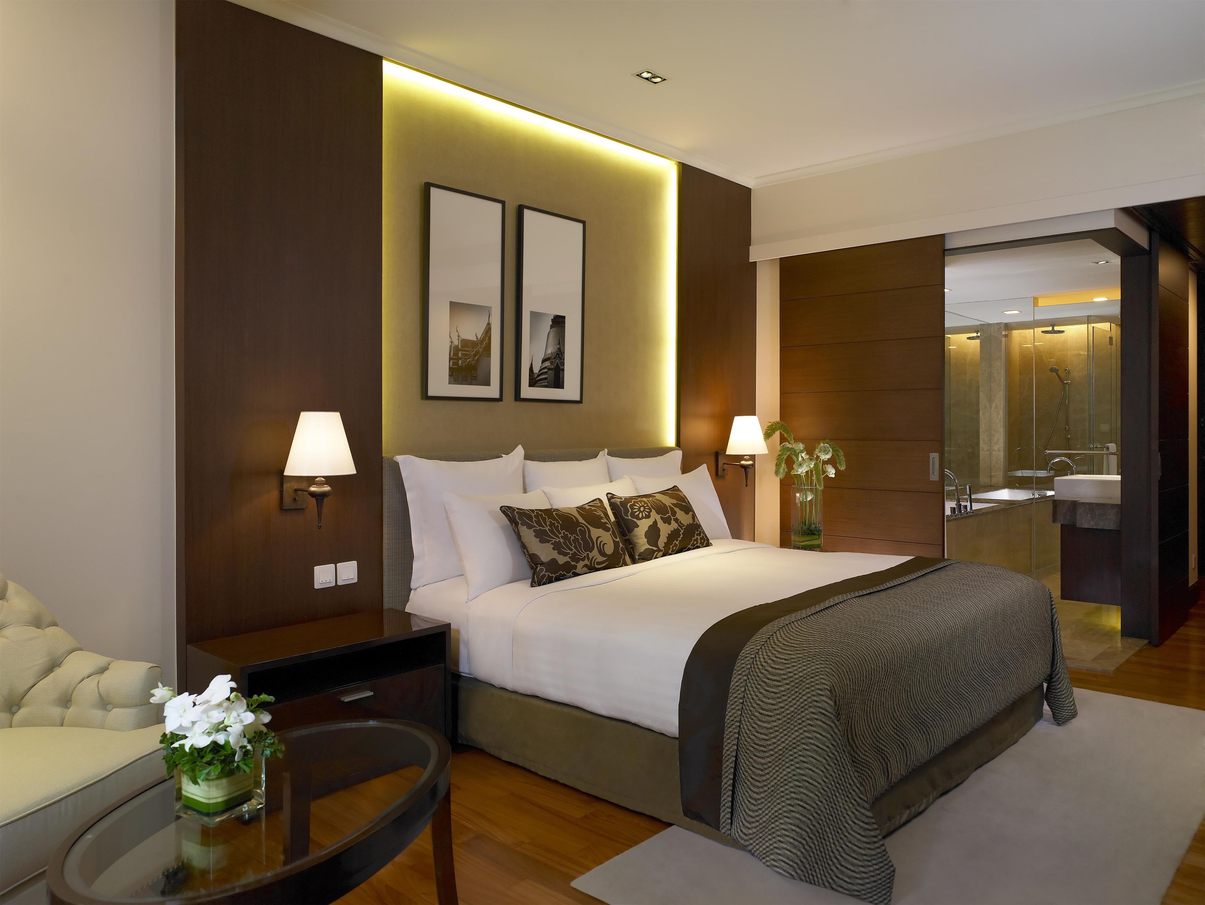 Una habitación de hotel moderna con una cama grande, lámparas de noche, una mesita con un florero y un baño con ducha y lavamanos.