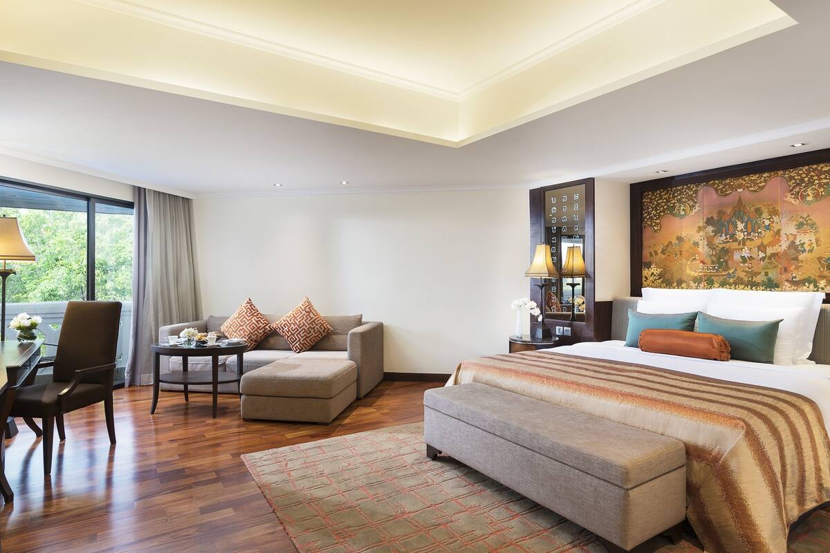 A hotel room with a large bed, a sofa, a small table, and a chair. There is a window with curtains, a lamp, and a decorative wall piece.