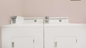 Deux machines à laver blanches à chargement frontal, dotées d’un panneau de commande et d’une pancarte au-dessus indiquant : « Savon et eau de Javel disponibles à la réception » et « L’hôtel n’est pas responsable des objets perdus ou volés ».