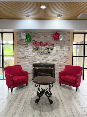 A room with a stone wall, a fireplace, two red armchairs, and a small round table. - HomeTowne Studios by Red Roof Birmingham - Highway 280 (Birmingham)