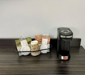 A coffee maker and a basket with various items including mugs and packaged goods on a wooden surface.