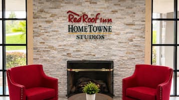 Red Roof Inn HOME TOWNE STUDIOS -hotellin aula, jossa on kiviseinä, kaksi punaista nojatuolia, takka ja pyöreä pöytä, jonka päällä on kasvi.