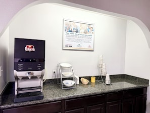 Estação de café com máquina de café Folgers, dispensador de água e um aviso sobre comodidades para estadias prolongadas.