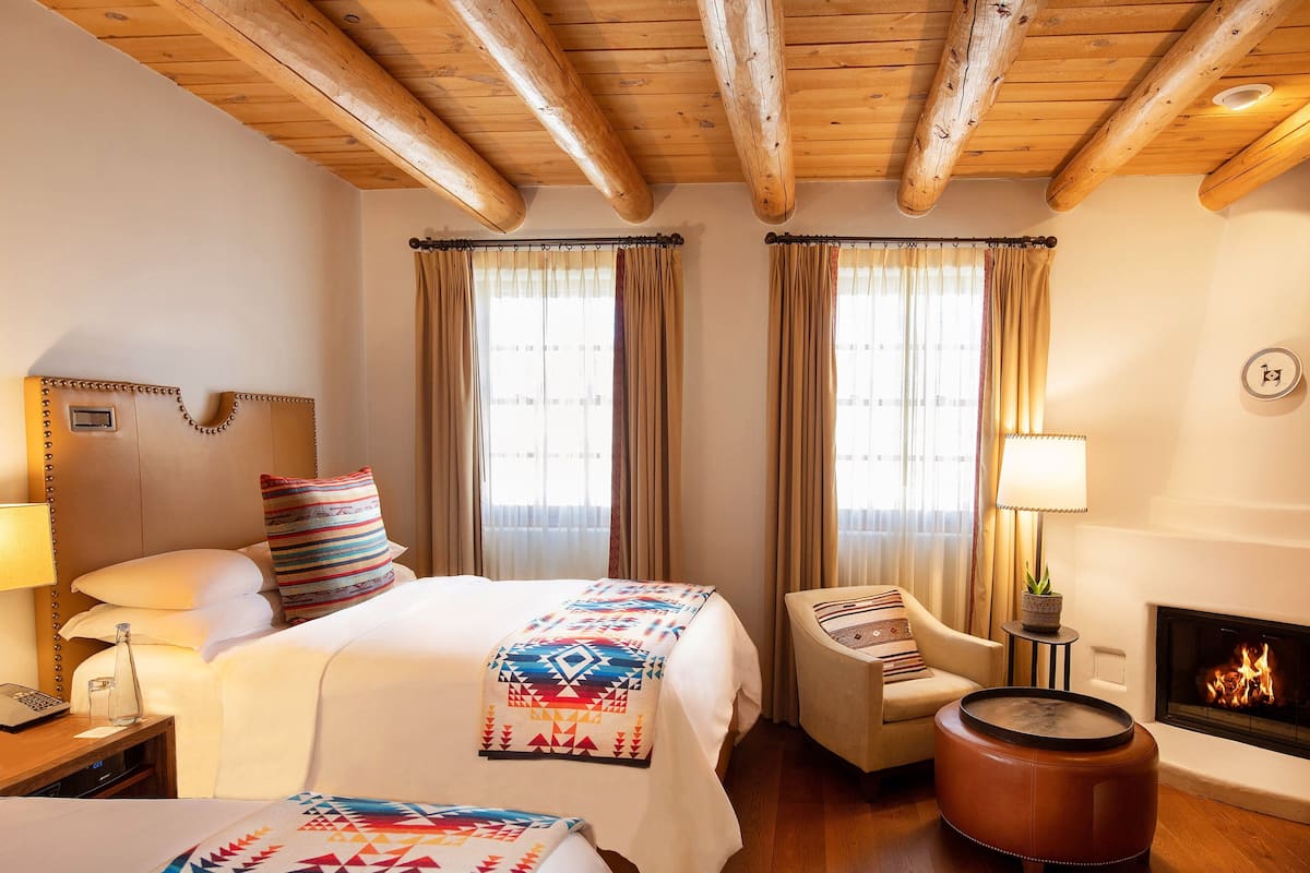 Habitación de hotel con techo de madera, una cama con manta de colores, una silla, una mesita con una planta y una chimenea.