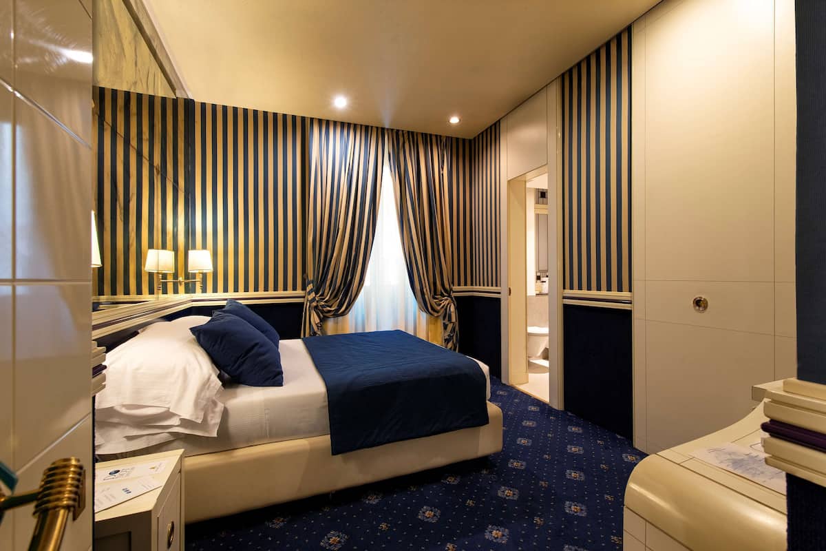 A hotel room with a bed, bedside tables, a mirror, curtains, and a patterned carpet.