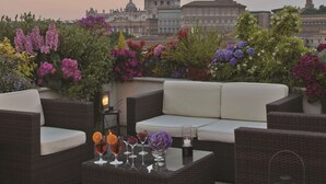 Une terrasse sur le toit, meublée de mobilier en osier, avec une table en verre où sont disposées des boissons, et une vue sur un bâtiment historique surmonté d’un dôme.