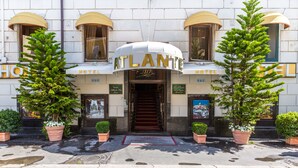 L’extérieur de l’HÔTEL ATLANTIS présente un style architectural classique, avec une entrée imposante, de grands pots de plantes et un tapis rouge menant à la porte.