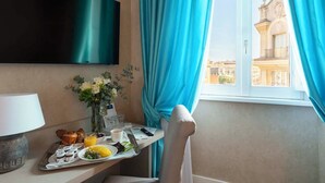 Une chambre d’hôtel dotée d’un téléviseur à écran plat fixé au mur, d’un plateau de petit-déjeuner avec des croissants, du jus d’orange et une assiette de nourriture, d’un vase rempli de fleurs et d’une vue sur les bâtiments à travers une fenêtre ornée de rideaux bleus.