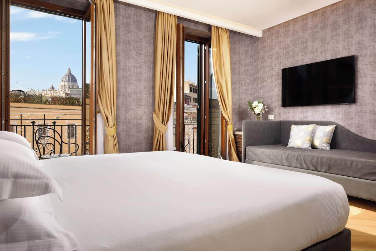 A hotel room with a large bed, a sofa, a TV mounted on the wall, and a balcony with curtains.