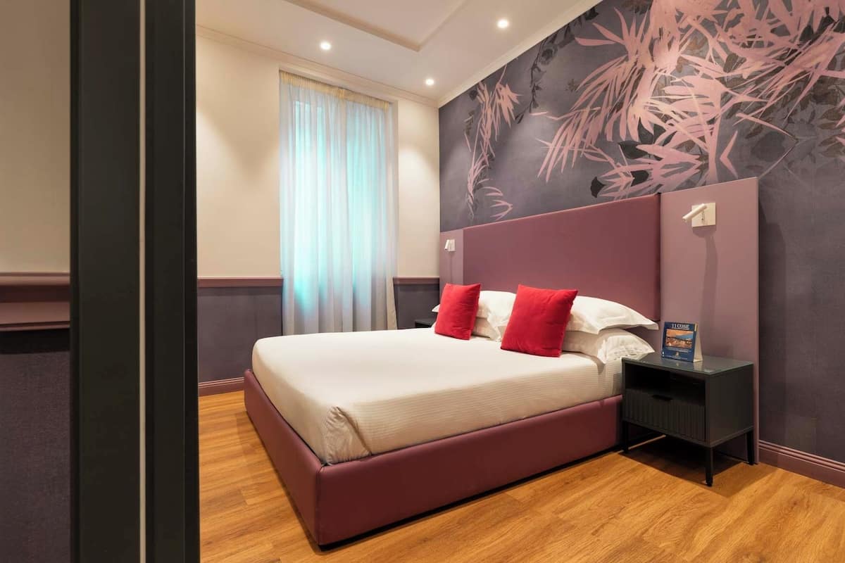 A modern hotel room with a large bed, red pillows, a nightstand, and a floral wall mural.