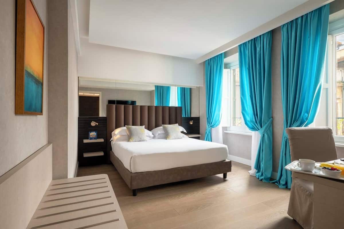 A modern hotel room with a large bed, a bedside table with a lamp, a chair, and a window with curtains.