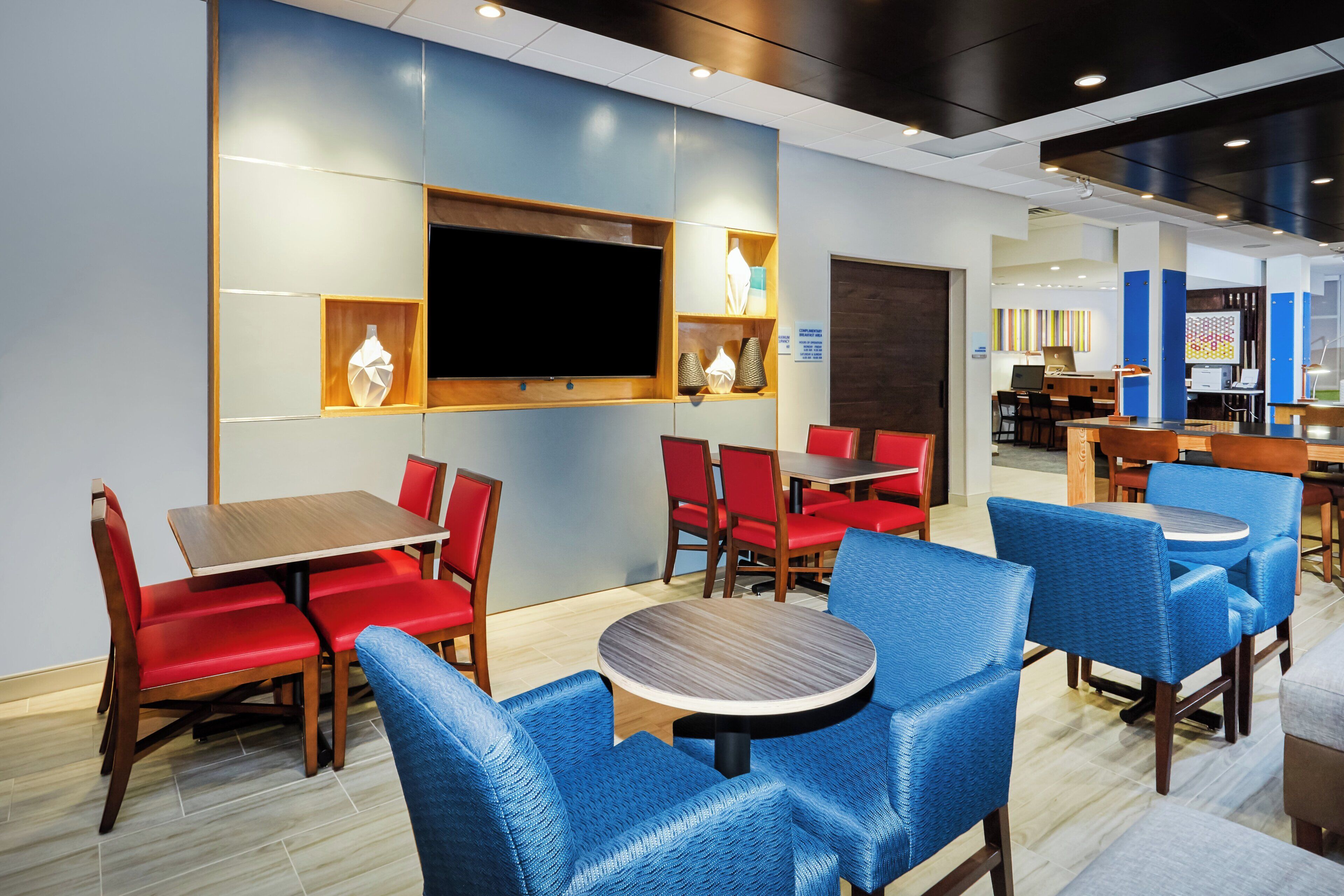 A modern hotel lobby with a flat-screen TV mounted on the wall, red and blue upholstered chairs, and a wooden reception desk.