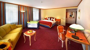 A hotel room with a bed, a sofa, a desk, a chair, a small table with fruit, and a TV.