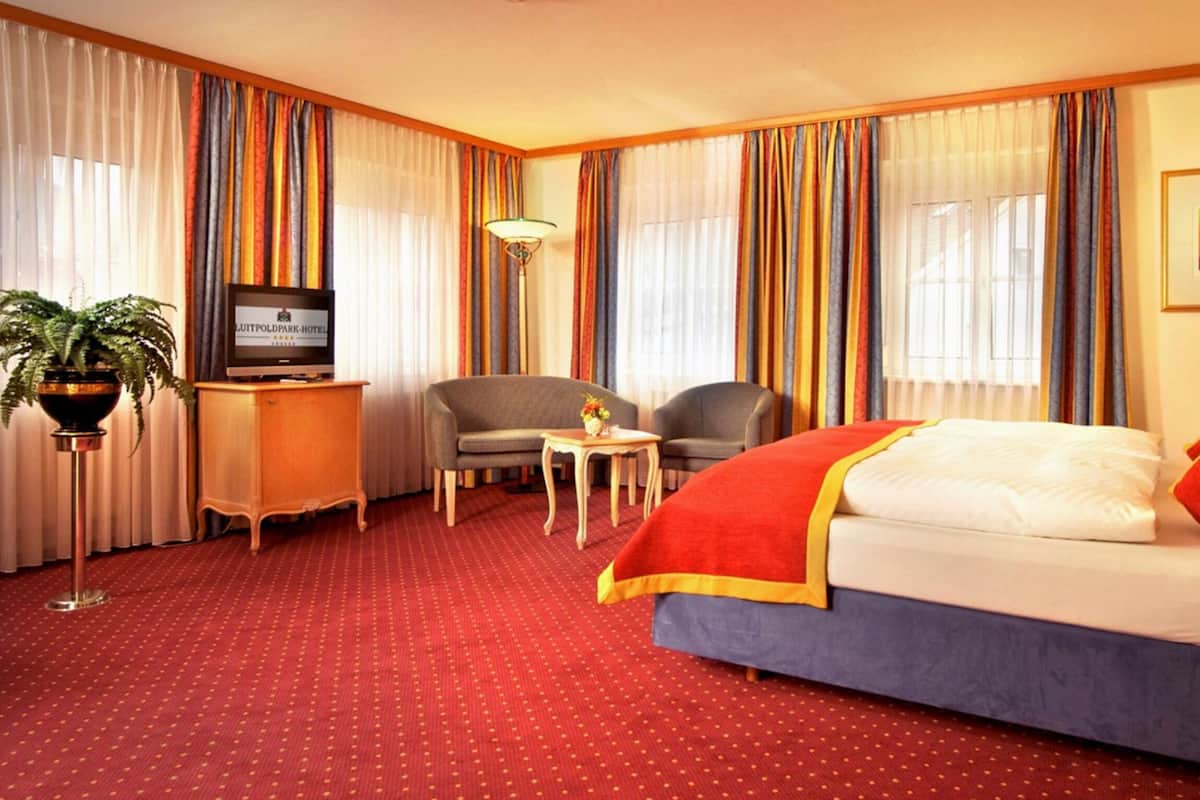 Quarto de hotel com uma cama, duas poltronas, uma mesinha, televisão e um vaso de planta.