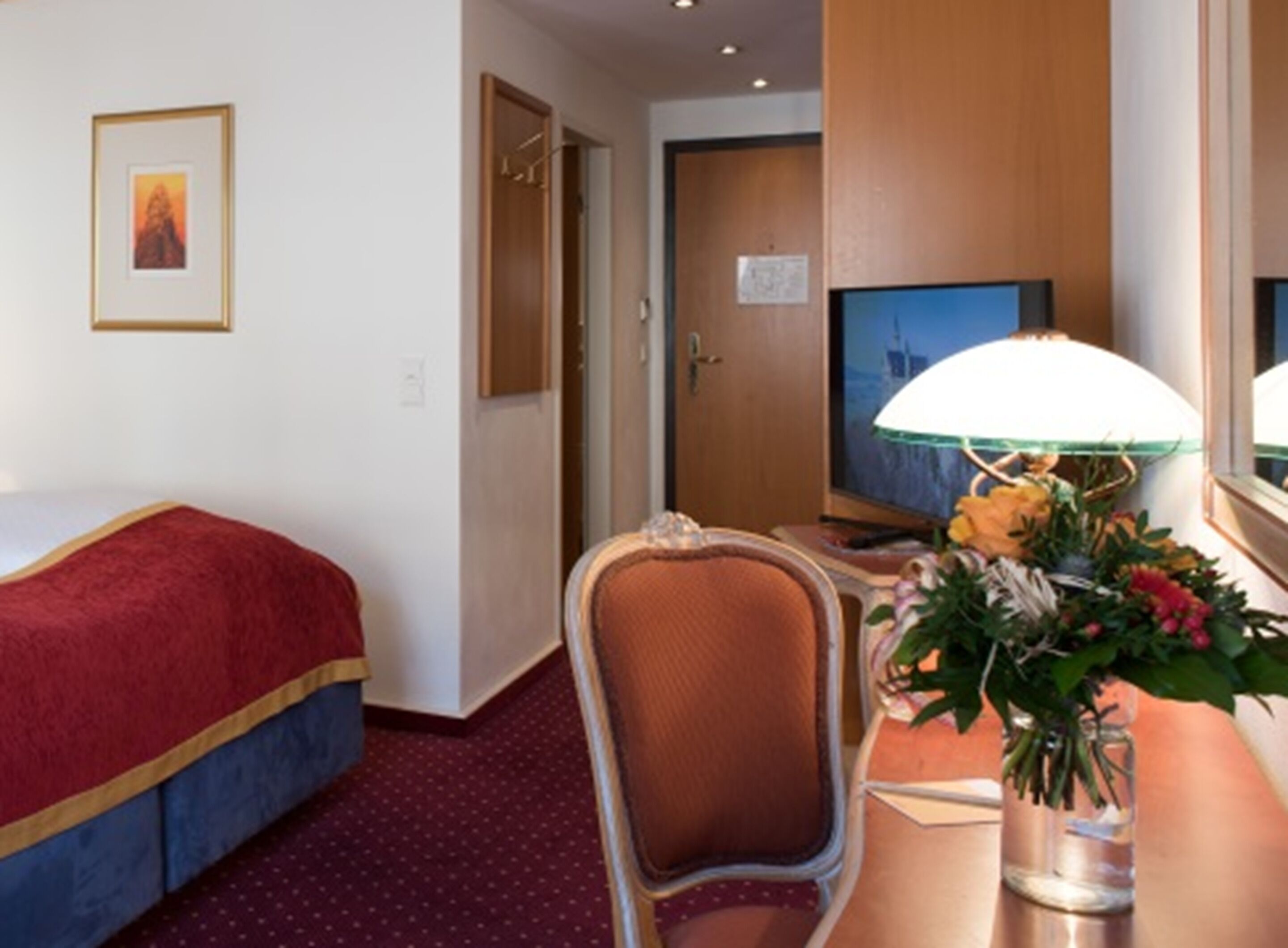 Una habitación de hotel con una cama, un escritorio con lámpara, una silla, un televisor y una puerta.
