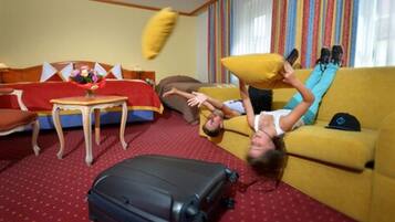 Habitación de hotel con un sofá rojo, un sillón amarillo, una mesa de centro de madera y una maleta negra en el suelo.