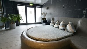 Un espace circulaire aménagé avec un banc rembourré et un sol carrelé, entouré de murs sombres et agrémenté d’une plante en pot.