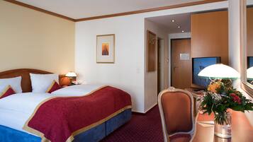 A hotel room with a large bed, a desk with a lamp, a chair, a television, and a framed picture on the wall.