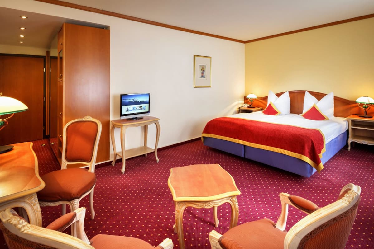 Quarto de hotel com uma cama grande, duas mesas de cabeceira com luminárias, uma televisão sobre uma mesinha e duas poltronas.
