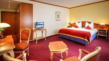 Een hotelkamer met een groot bed, twee nachtkastjes met lampen, een televisie op een bijzettafeltje en twee fauteuils.