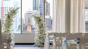 Pasillo con sillas blancas y decoración floral, que conduce a una ventana con vista panorámica de la ciudad.