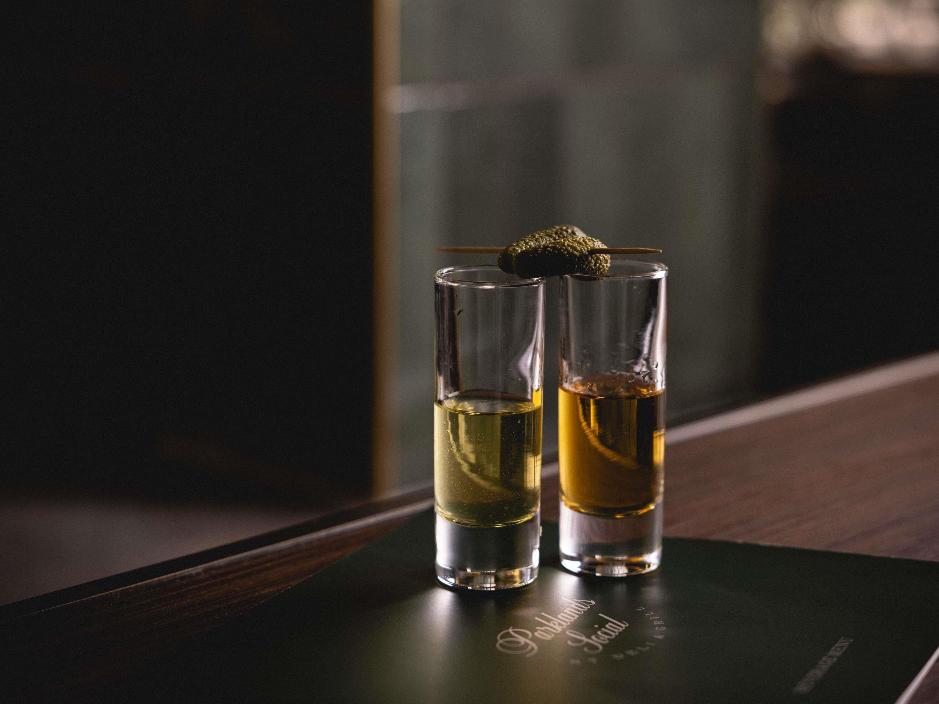 Dos vasos para shots sobre una superficie oscura con un logo.