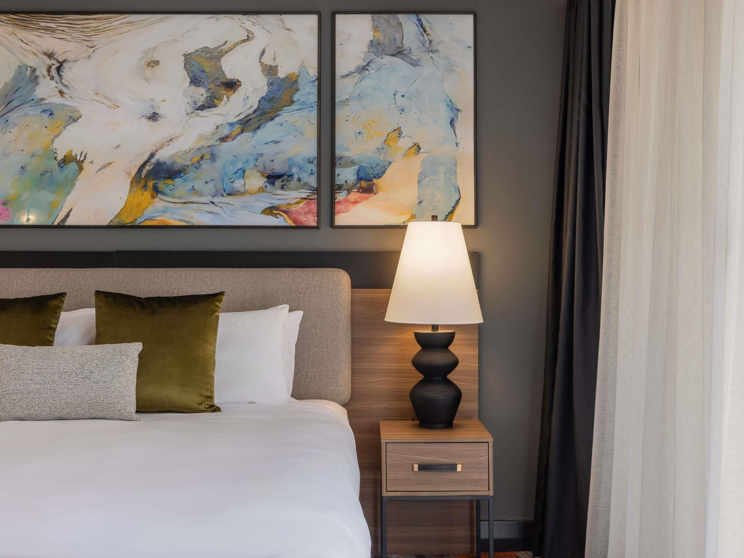Habitación de hotel con cama, mesita de noche con lámpara y arte abstracto en la pared.