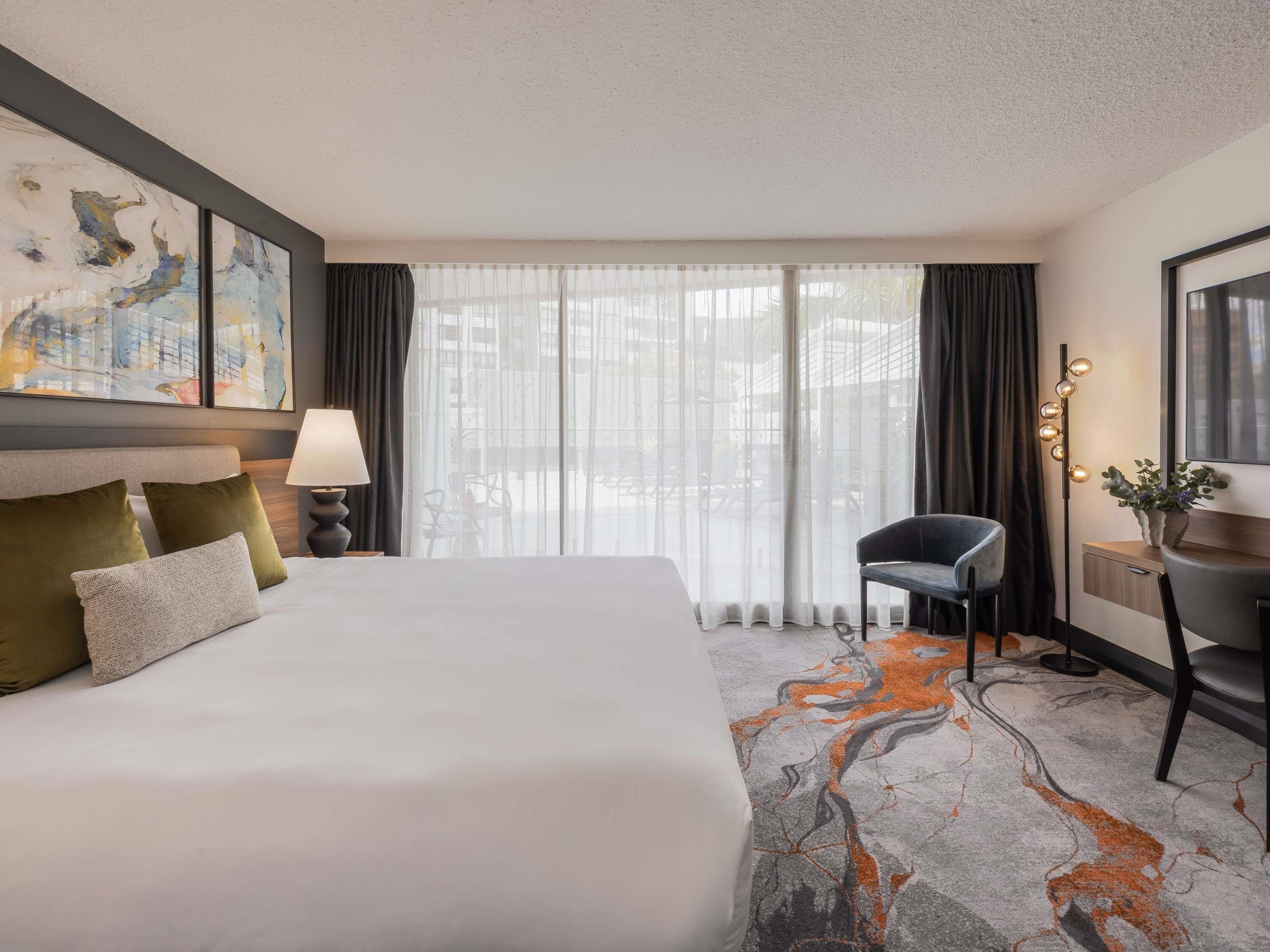 A hotel room with a bed, a desk, a chair, a lamp, a painting, and a view through the window.