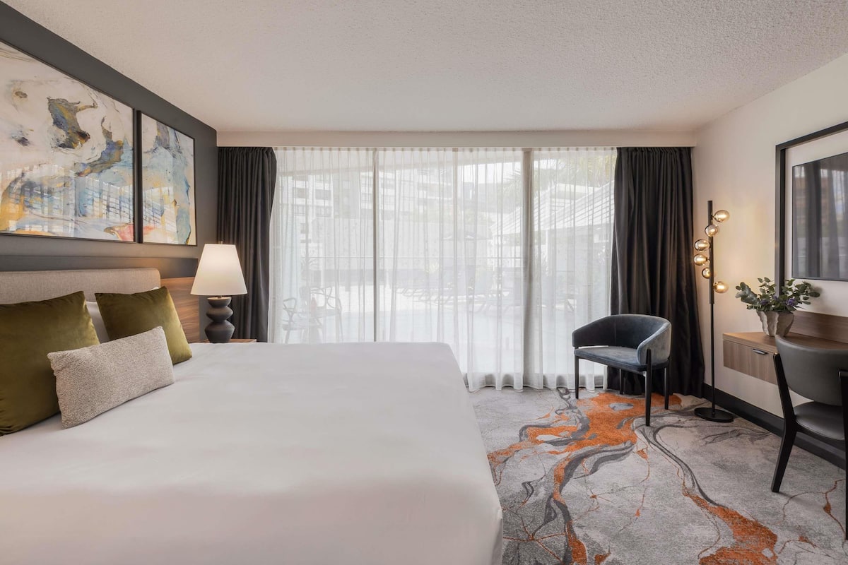 A hotel room with a bed, a desk, a chair, a lamp, a painting, and a view through the window.