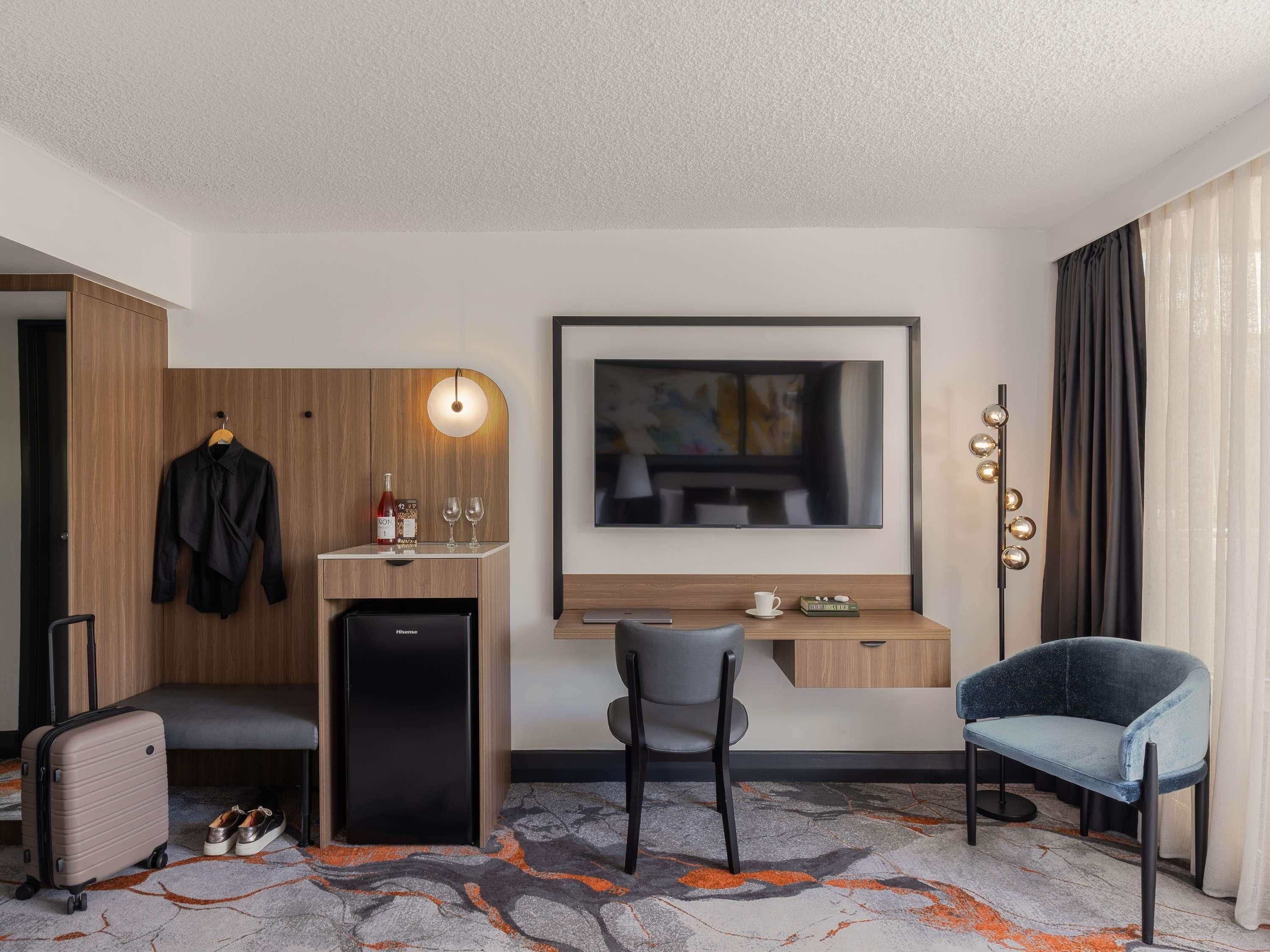 A hotel room with a desk, chair, minibar, and a TV mounted on the wall.