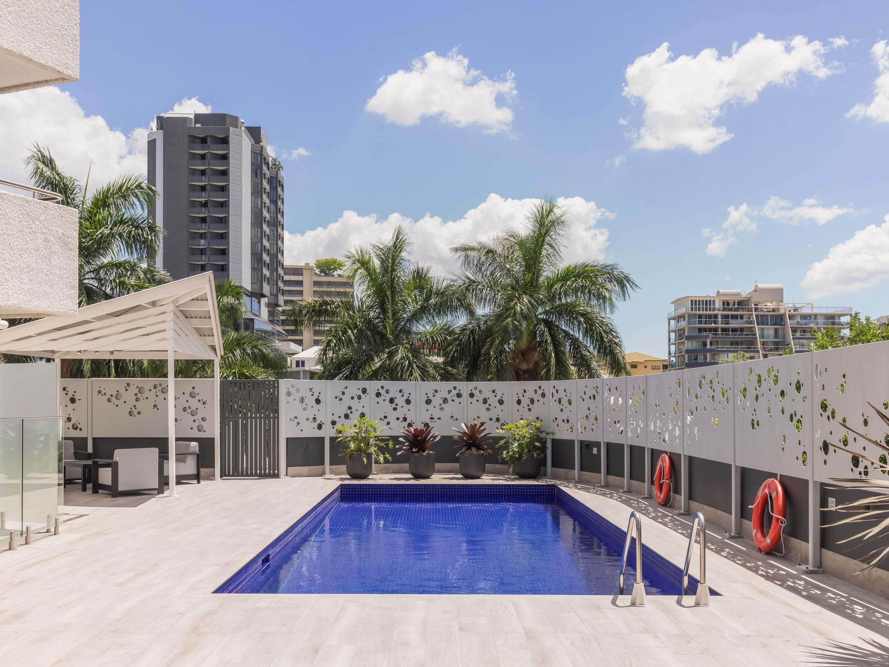 A rooftop pool with a blue pool, surrounded by potted plants, a white fence with circular cutouts, and red lifebuoys.