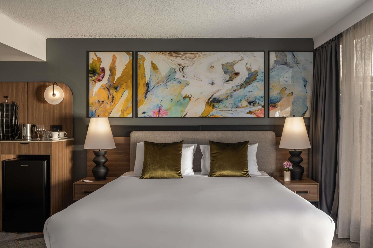 A hotel room with a large bed, two bedside lamps, a coffee machine, and abstract art on the wall.