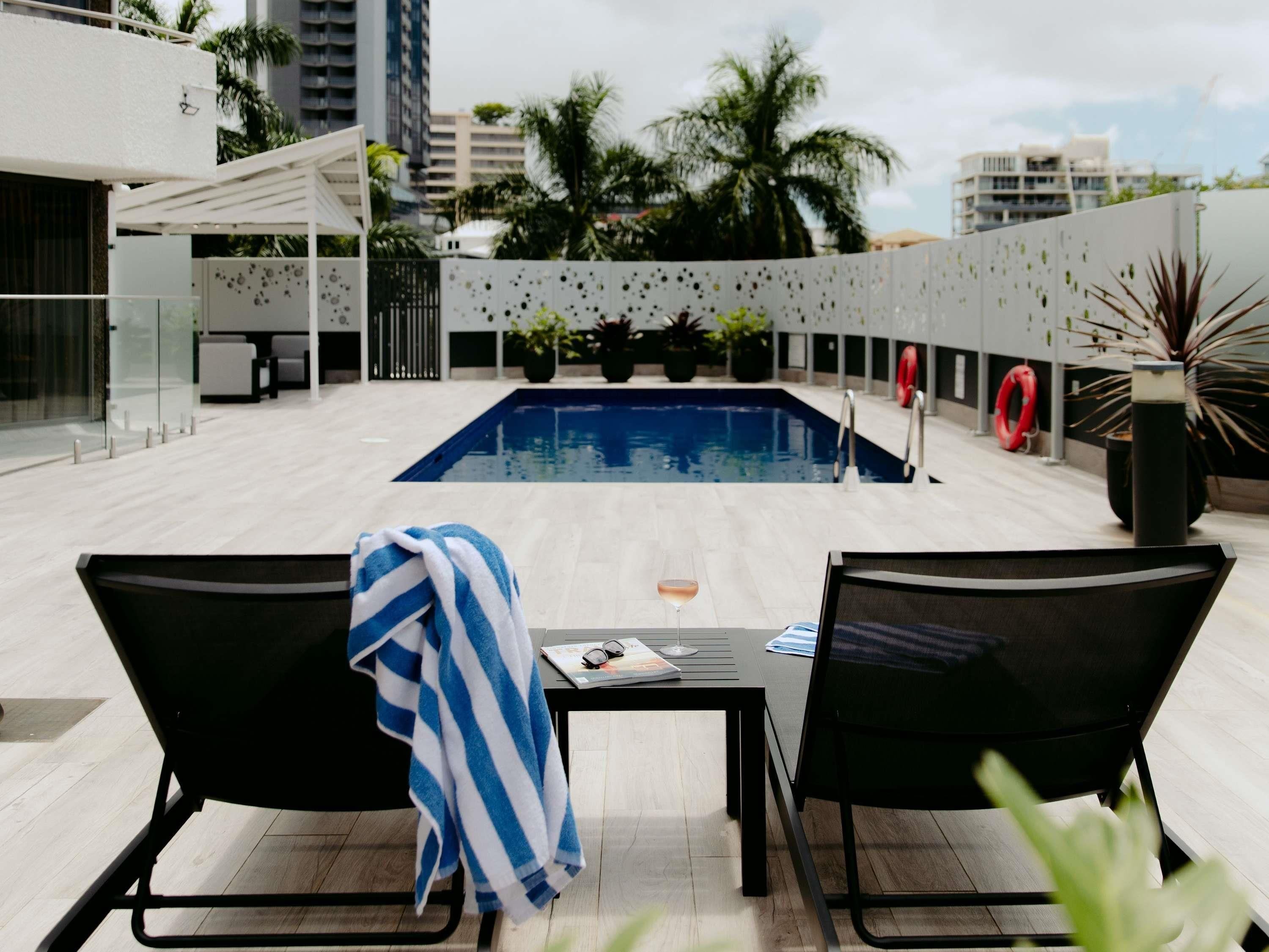 A rooftop pool area with two lounge chairs, a table, and a view of the cityscape.