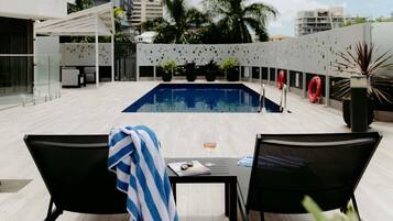 A rooftop pool area with two lounge chairs, a table, and a view of the cityscape.