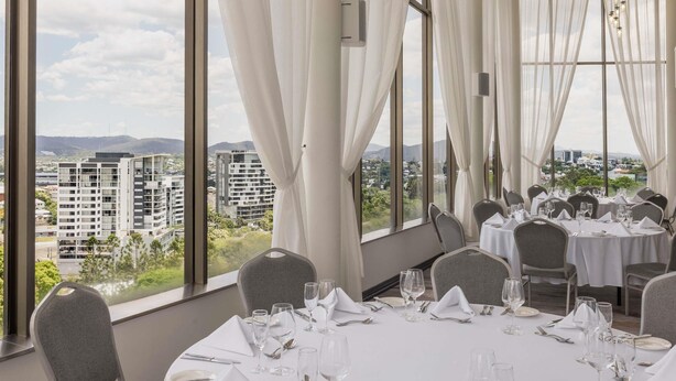 Un comedor con mesas dispuestas para comer, ventanas amplias con cortinas y vistas a edificios y follaje.