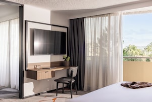 Une chambre d’hôtel comprenant un lit, un bureau avec une chaise, une télévision et une fenêtre avec des rideaux.