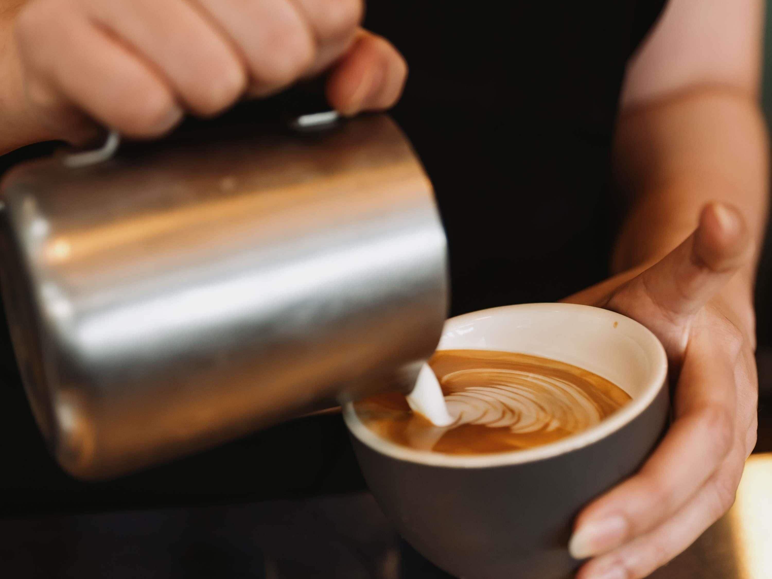 Iemand giet melk in een kop koffie, waardoor een latte art patroon ontstaat.